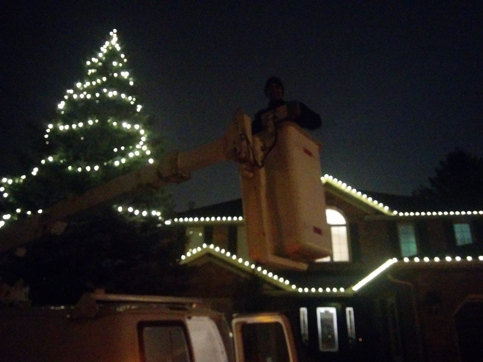 Worker in an aerial lift bucket installing warm white roofline Christmas lights on a two-storey brick home.