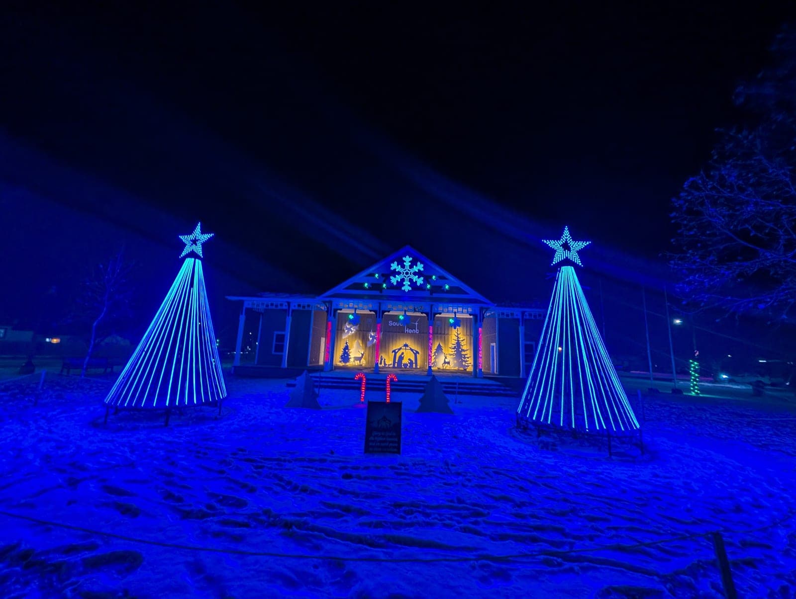 Community pavilion holiday display with two blue spiral light-cone trees, lit stars and a nativity scene on fresh snow at night.