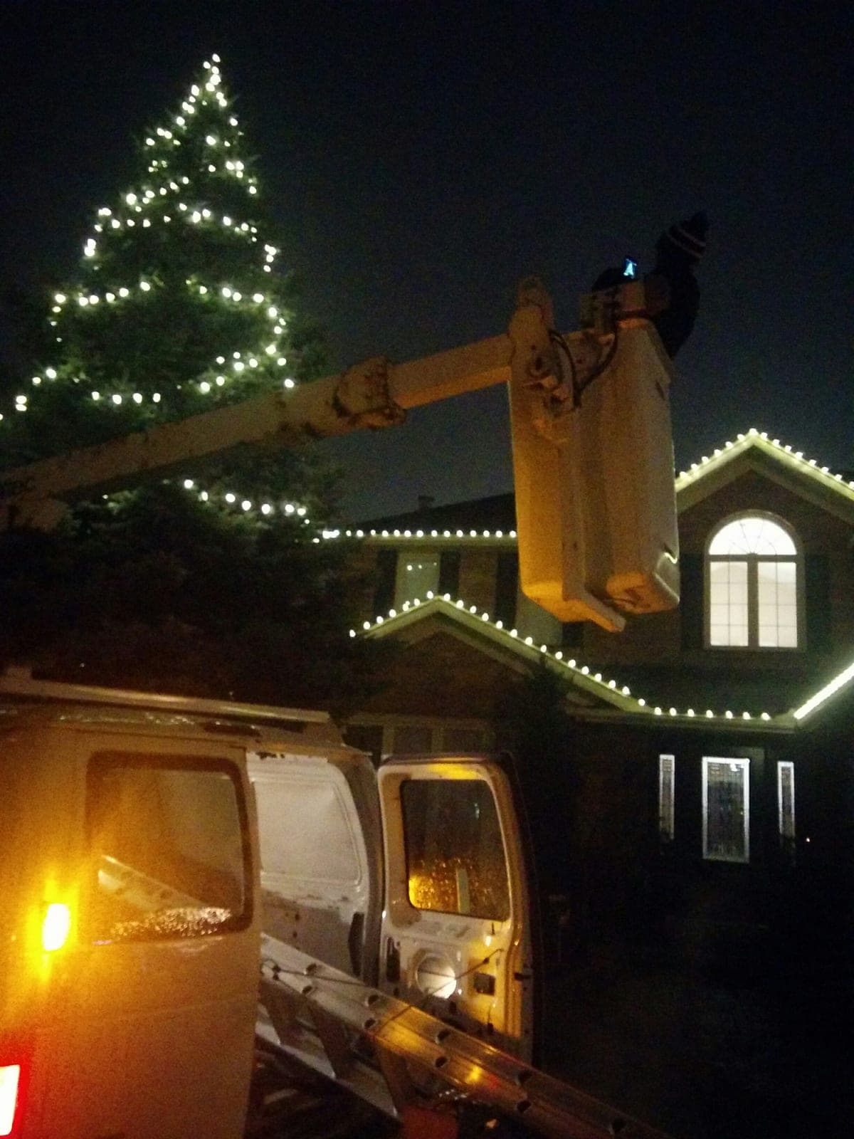 Christmas light installer in a bucket lift truck hanging warm white roofline lights on a two-storey home at night.