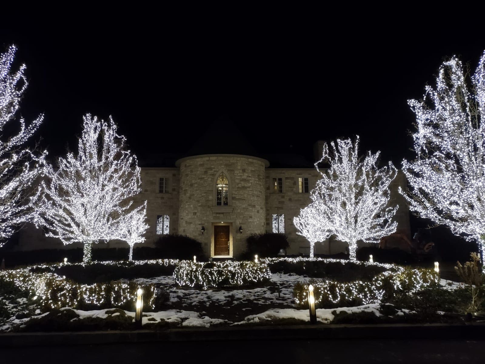Stone estate with trees wrapped in brilliant cool-white Christmas lights and lit garden hedges in snow