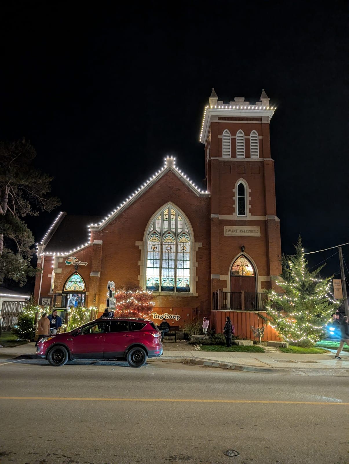 Historic brick church outlined in warm white roofline lights with light-wrapped evergreen trees at night.