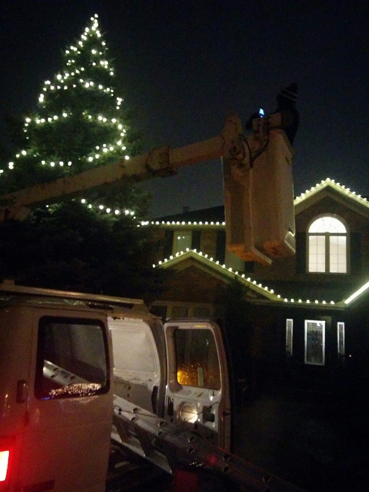 Holiday lighting technician in a bucket truck installing Christmas lights on a two-storey home and evergreen tree at night