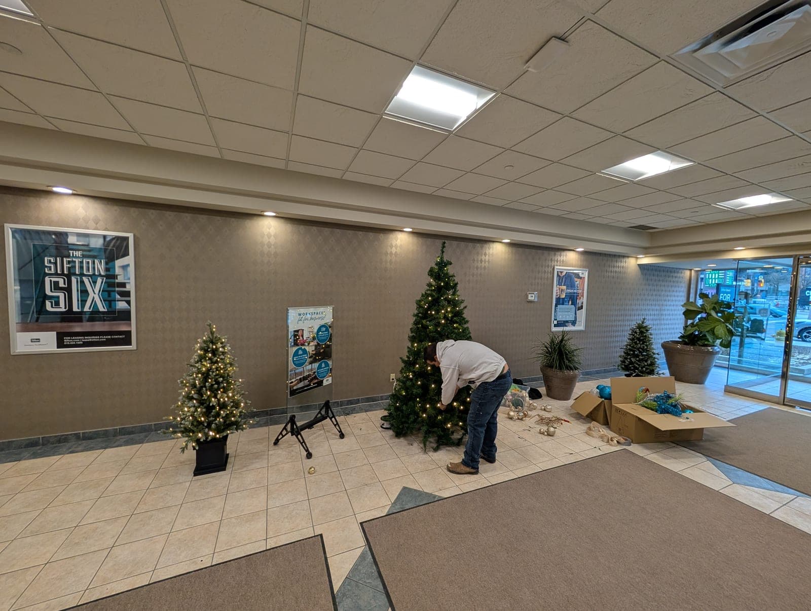 Christmas decor installer trimming a lit tree in an office building lobby.