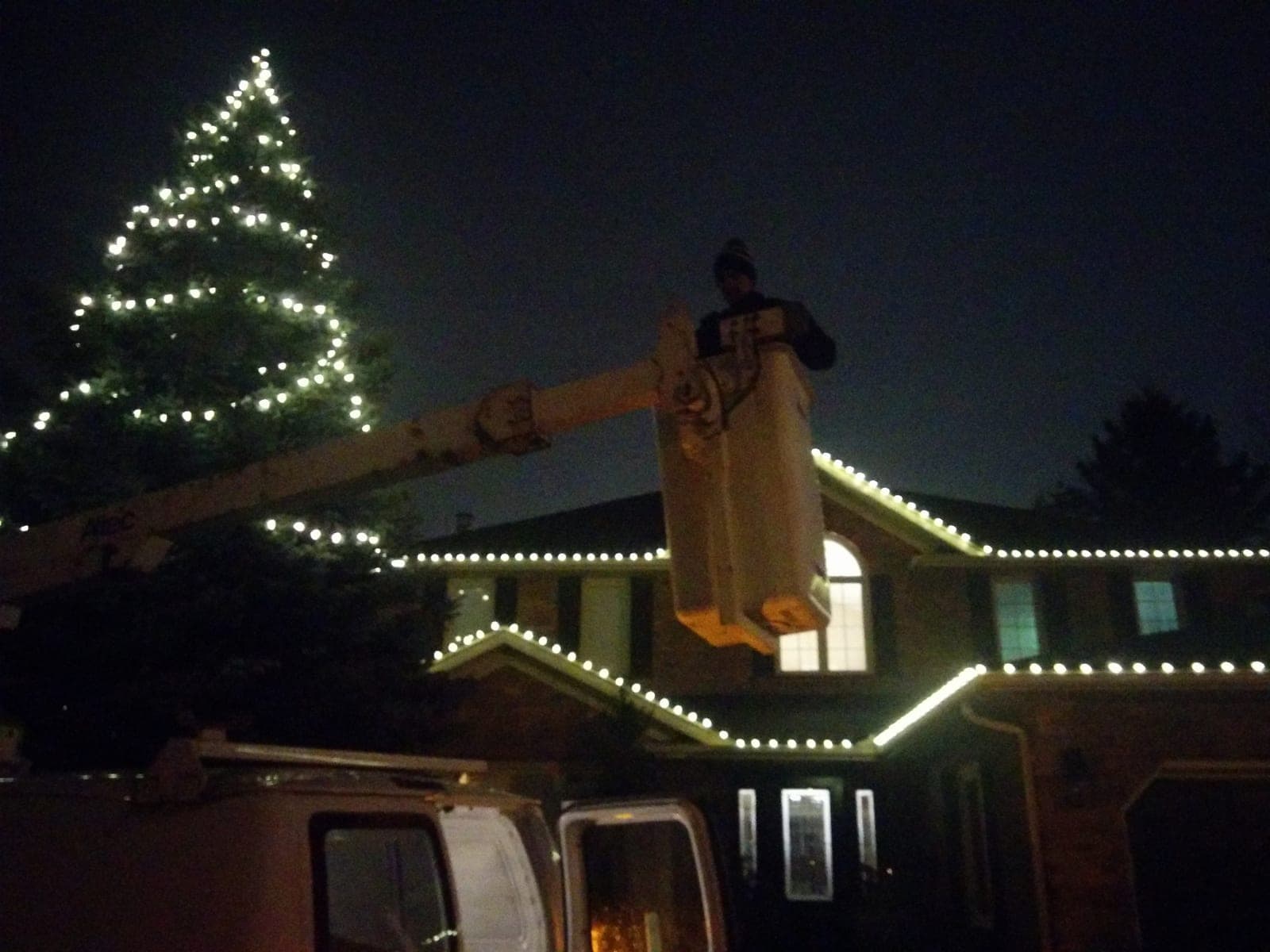 Christmas light installer working from an aerial bucket to hang warm white lights on a brick home's roofline.