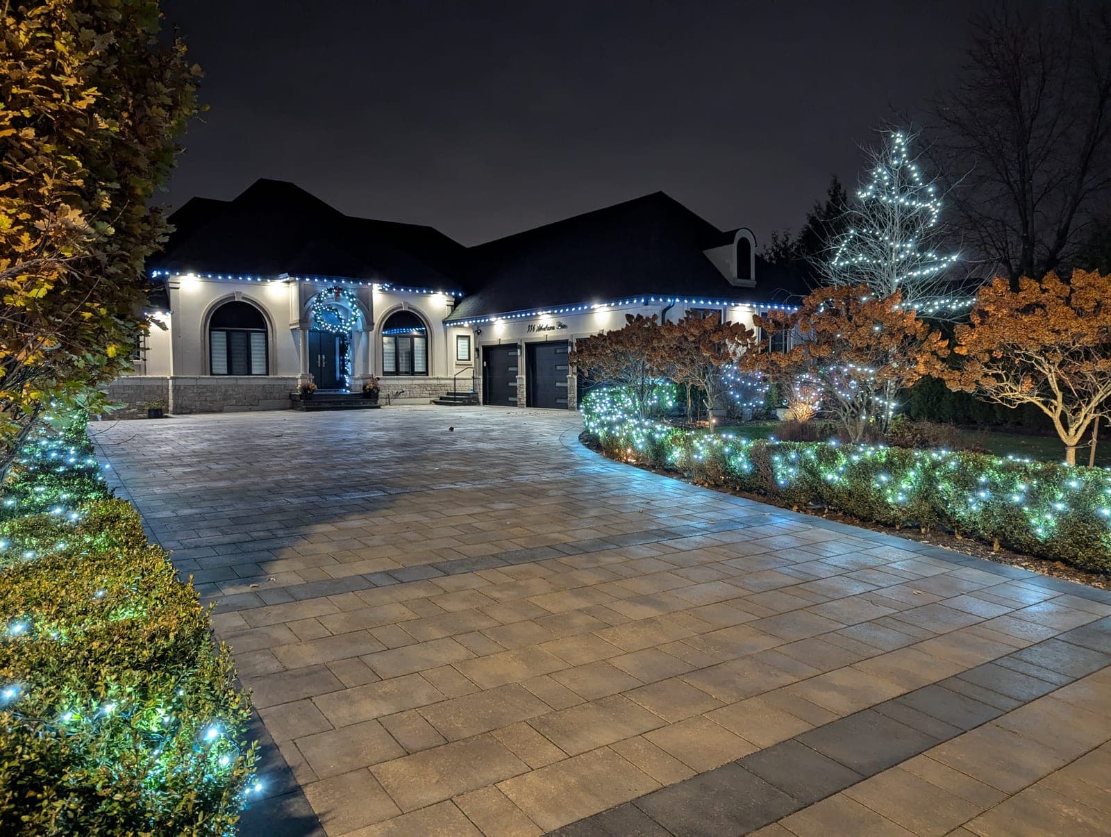 Luxury home with roofline Christmas lights and cool-white lit garden beds along a paver driveway