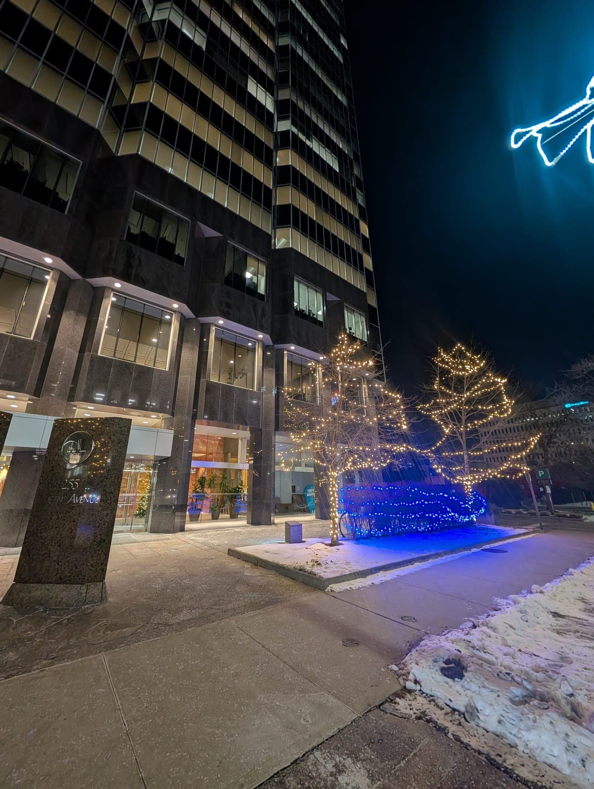 Office tower plaza with warm-white tree wraps, blue-lit shrubs and an illuminated angel light display.