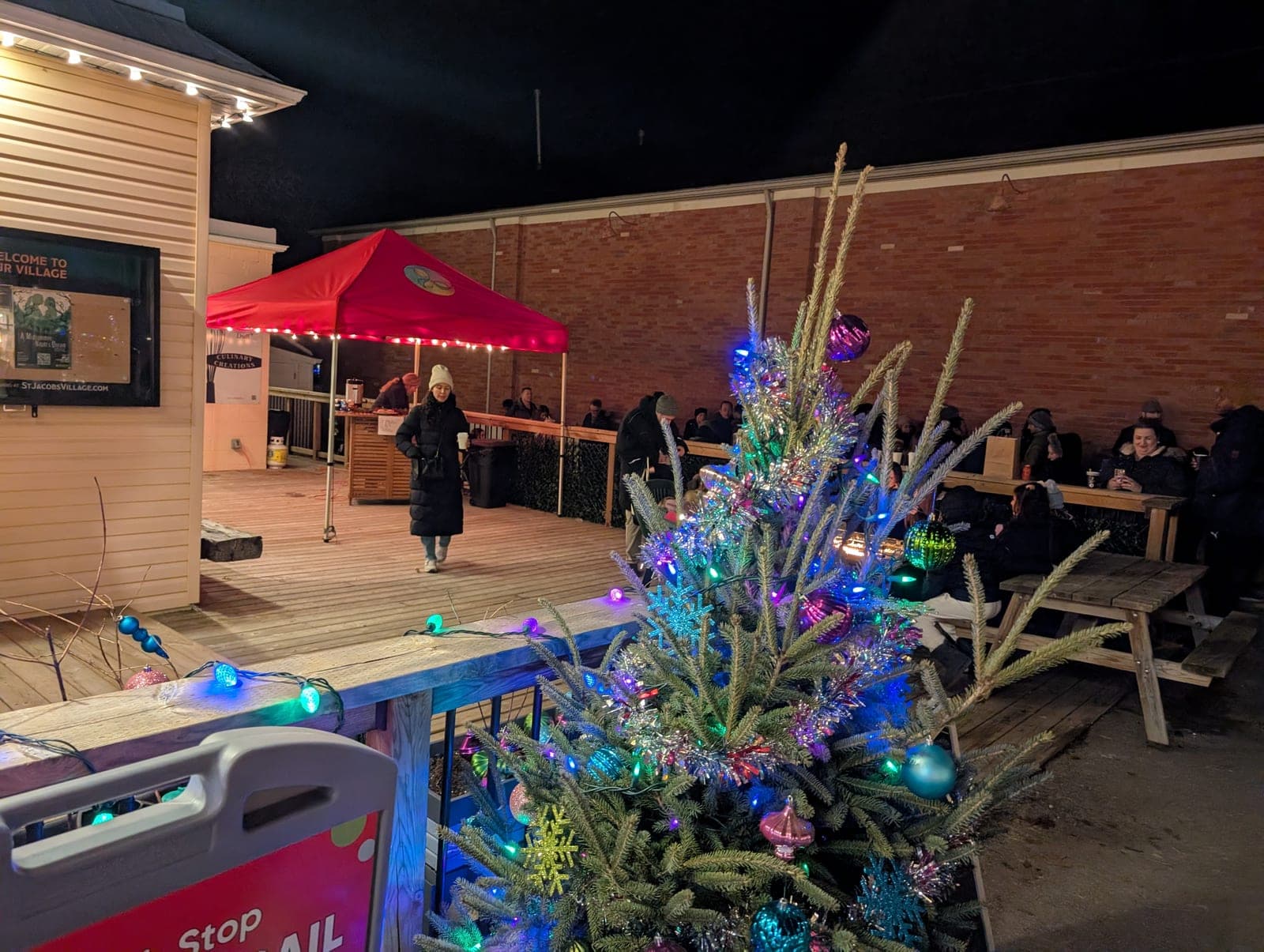 Brightly decorated outdoor Christmas tree on a market patio with string lights and an evening crowd.