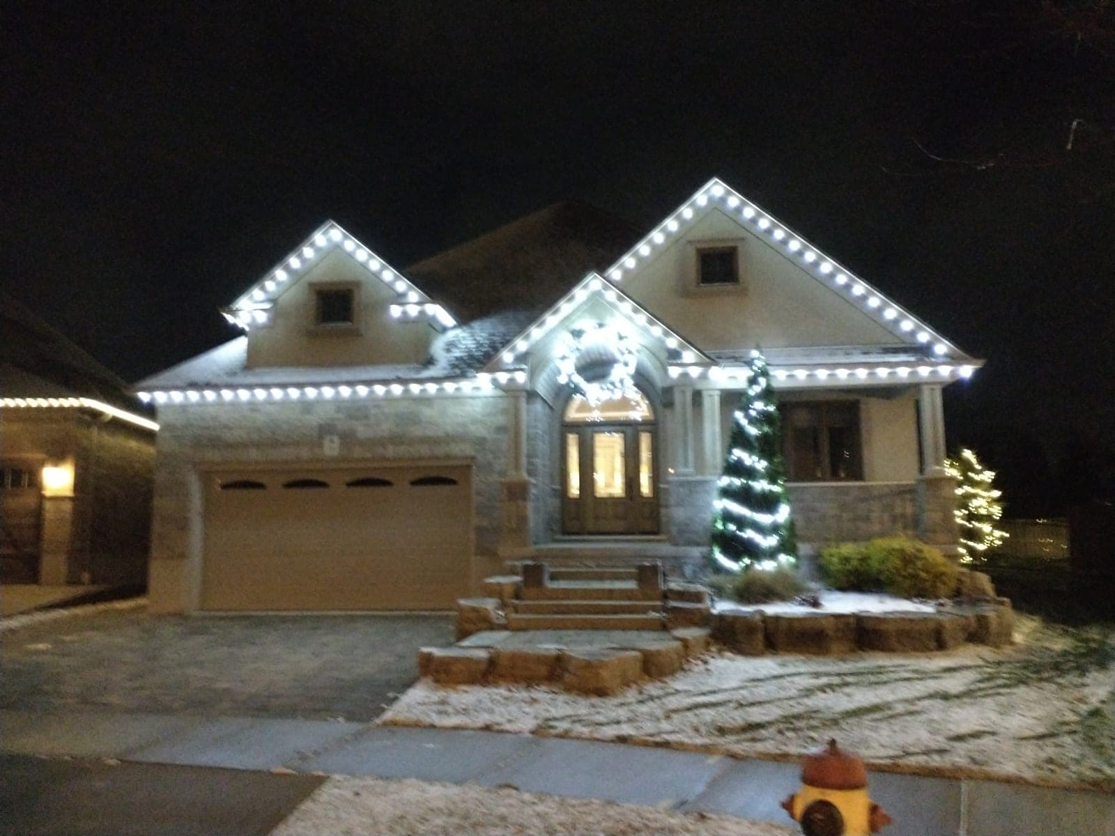 Home with warm-white roofline Christmas lights, an illuminated wreath and a lit evergreen by the porch
