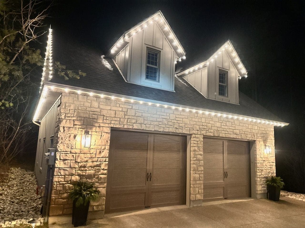 Two-storey stone coach house outlined in warm-white roofline Christmas lights tracing dormers and eaves at night