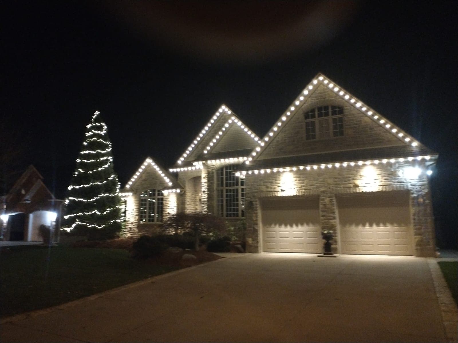 Large stone home outlined in warm-white roofline Christmas lights beside a tall lit spruce tree