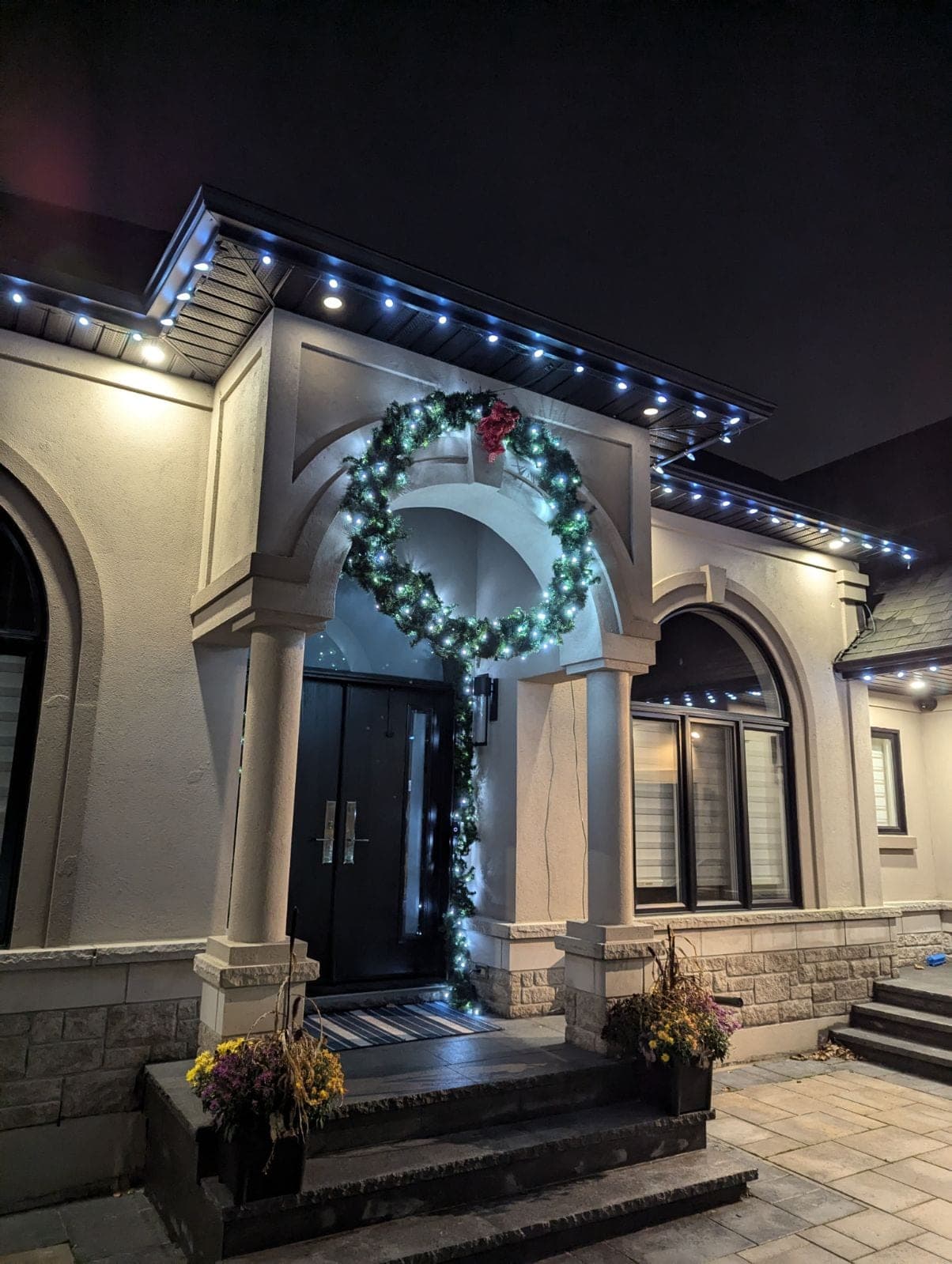 Home front entrance with warm-white roofline and soffit lights, pot lights and a lit garland-wrapped wreath at night