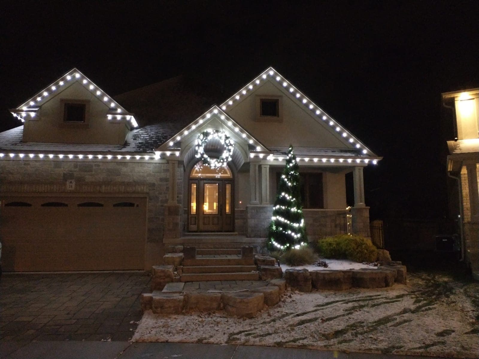 Stone bungalow front entry outlined in white C9 roofline lights with a lit wreath and cone tree.